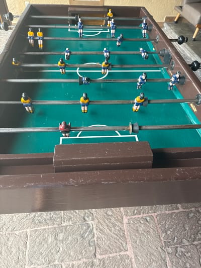This media file captures a close-up, angled view of a vintage or well-used foosball table, set in what appears to be an indoor or covered outdoor area in Tres de Mayo, Mexico. The image is oriented vertically (rotated 90 degrees counter-clockwise), but if viewed upright, the foosball table extends from left to right.
The primary subject is the foosball table itself, which features a dark brown wooden frame showing signs of wear and tear, including scuffs and dust. The playing surface is a distinct green, marked with white lines delineating the field and goal areas. Metal rods run across the table, each equipped with several small, plastic player figurines. Most players are dressed in either blue and yellow jerseys, representing two teams, while one player figurine, positioned closer to the right goal, is notably red. Black handles are visible at the ends of some rods outside the table's frame.
No active game is taking place; the players are stationary on their rods. The lighting is diffused, suggesting an indoor setting or a shaded outdoor space, and does not indicate a specific time of day. The table rests on a textured, light-colored stone-paved floor, characterized by irregularly shaped stones and visible grout lines, hinting at traditional architecture often found in Mexico. A lighter-colored wall or structure, possibly with wooden accents, is faintly visible in the background to the left. No visible text is present in the image.
