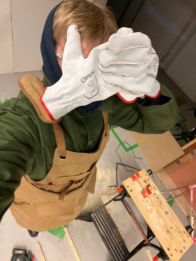 From a high-angle view, a person wearing a green jacket, blue beanie, and tan canvas apron holds up gloved hands in front of their face. The white gloves feature red trim and the brand "OPSIAL." In the background, a workbench with a wooden top and black frame is visible. Other elements include a pink and white bicycle frame, scattered wood scraps, and a concrete floor with green lines.