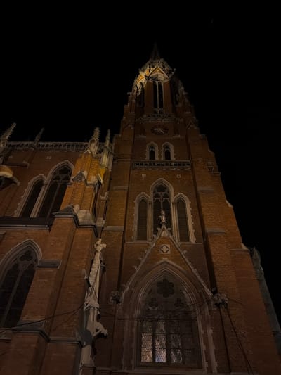 The image depicts the Church of St. Peter and Paul in Osijek, Croatia, at night. The church's architecture is prominently displayed, showcasing its brick construction, arched windows, and ornamental stonework. A clock is visible on the facade. The scene is dimly lit, highlighting the architectural details against the dark sky. No people are visible in the frame.
