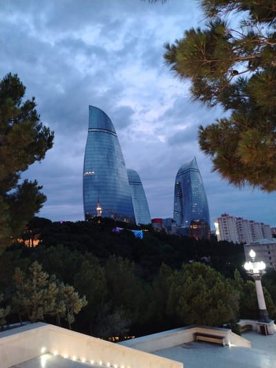 This media file depicts the Flame Towers in Baku, Azerbaijan, at dusk. The three flame-shaped skyscrapers, clad in glass, dominate the skyline. The building on the left is partially obscured by trees and the building in the center is the most prominent. The third building is to the right of the center building and is also visible.

The foreground features a stone walkway with embedded lights and a bench. Trees and lush greenery create a natural frame around the architectural marvels. A vintage lamppost stands to the right, its multiple lights casting a warm glow. In the mid-ground, dense foliage covers a hillside, with some structures and colorful lights visible amongst the trees. The sky is overcast, with shades of gray and blue, suggesting a dramatic evening atmosphere. The overall scene evokes a sense of urban grandeur and natural beauty, with the setting sun casting a soft light on the cityscape. The time of day is indicated by the twilight sky and the illumination of artificial lights. No people are visible in the frame.
