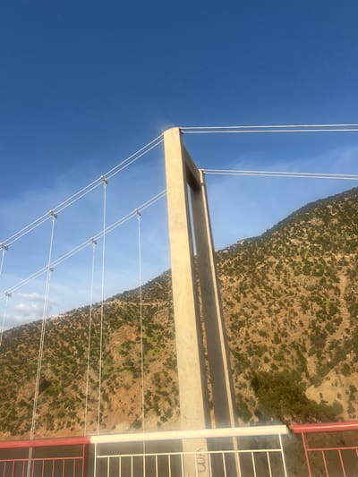 Captured from an elevated perspective, the image presents a view of Ait Mazigh, Morocco. A bridge structure occupies the foreground, with a clear blue sky dominating the upper left quadrant. The land is characterized by rugged, mountainous terrain interspersed with vegetation. A section of a building with a red frame and white vertical supports is visible in the right portion of the frame. Electrical lines are present, spanning across the scene. The image suggests a rural or semi-rural setting, typical of the terrain in the Ait Mazigh region.