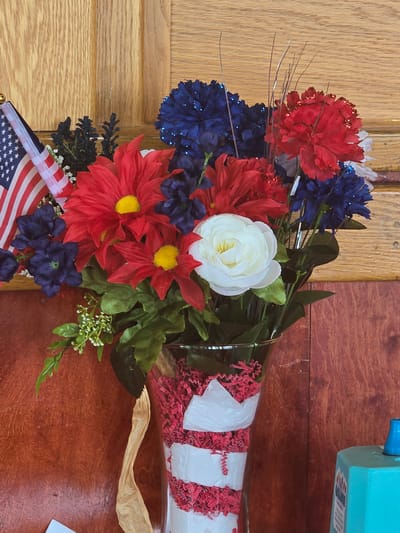 A floral arrangement with American flags is displayed in a glass vase decorated with red and white stripes, sitting on a brown surface against a wooden background. The arrangement features red and blue artificial flowers, including daisies and carnations, with a white rose in the center. A small American flag on a pole is positioned to the left of the flowers. The vase is filled with red shredded paper. The background consists of two wooden panels, one lighter and one darker, suggesting a room interior. The lower right corner shows a portion of a blue object, possibly a dispenser or container. The scene appears to be set up for a patriotic celebration or event. The image was captured indoors, likely in Cumming, Georgia, United States of America.