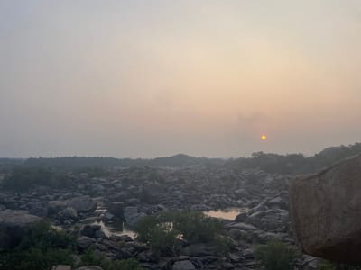 The image depicts a hazy, panoramic landscape in Hale Bandi Harlapura, India, at either sunrise or sunset. The scene is dominated by a vast, rocky terrain under a diffused, pale sky. The upper two-thirds of the frame show a sky graduating from light grey-blue on the left to a soft orange-yellow on the right, where a distinct, yellow-orange sun is visible as a low disc, partially obscured by atmospheric haze.
The lower portion of the image features a sprawling expanse of dark grey, smooth boulders and rocks. A narrow, reflective body of water, likely a river or stream, winds through the center of this rocky landscape, capturing the pale light from the sky and sun. Sparse, low-lying green vegetation, consisting of shrubs and small trees, is interspersed among the rocks and along the water's edge. A large, light brown boulder is partially visible in the immediate lower-right foreground, adding depth. No people, animals, or artificial structures are discernible within the frame. The overall environment appears rugged and natural.