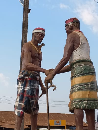 Two statues of men stand with clasped hands, a walking stick between them. They wear traditional attire, including red-topped caps. The man on the left is shirtless with a beaded necklace and a bird perched on his shoulder; he wears a plaid skirt. The man on the right wears a white tank top and a skirt with geometric designs. An MTN billboard sits below the statues. A utility pole is on the left side of the statues and the background features a bright blue sky.