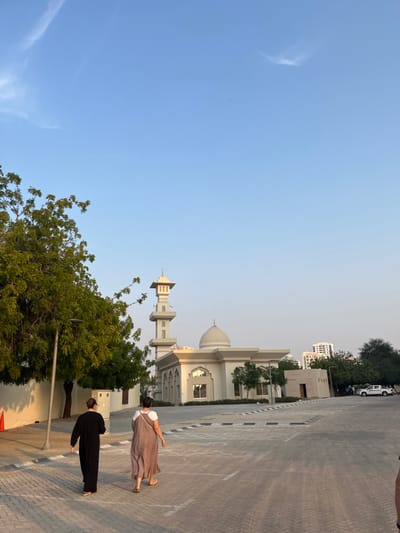 The image displays an outdoor scene in Sharjah, United Arab Emirates, under a clear, light blue sky with faint wispy clouds. The lighting indicates late afternoon or early evening, casting long shadows and illuminating structures with a warm, golden hue.
In the mid-ground, a light-colored mosque is the central architectural feature. It possesses a prominent central dome and a single, square-profile minaret composed of multiple tiered sections, topped with a finial. The mosque's facade includes arched window openings and decorative elements.
Two adult individuals are walking away from the camera on a paved surface, moving towards the mosque. The individual on the left wears a long, dark-colored garment. The individual on the right wears a loose-fitting, light brown or beige sleeveless dress. Both individuals have uncovered heads and wear flat-soled footwear. Their backs are largely presented to the viewer, and no direct interaction is discernible.
The ground consists of interlocking pavers, marked with white and black lines that delineate parking spaces or pathways. To the left, a large tree with dense green foliage partially obscures a dark-colored lamppost and a light-colored utility box situated against a continuous light-colored wall. An orange traffic cone rests near the base of the tree on the left.
In the background, beyond the mosque, several multi-story buildings rise on the horizon, confirming an urban setting. A white pickup truck is visible parked in the far right distance, partially obscured by trees. The environment appears calm and clear.