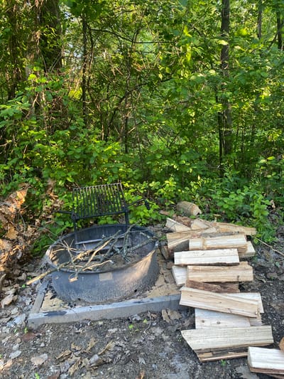 The image shows a fire pit in an outdoor setting. The fire pit is made of dark metal and is surrounded by a concrete base. There is a metal grill rack placed over the fire pit. A pile of firewood is stacked neatly next to the fire pit. The scene is surrounded by lush green foliage, suggesting a wooded or natural area. The ground is covered with dirt, fallen leaves, and some small plants. The lighting suggests it is daytime, with sunlight filtering through the leaves, creating dappled shadows. The overall impression is of a prepared spot for a campfire in a natural environment. There is no text visible in the image. The location is the Town of Peru, USA.