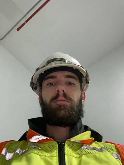 A man wearing a white hard hat and a lime green safety vest with reflective orange and silver stripes stares directly at the camera from an unknown location in Kempton Park, South Africa. He has short dark hair, brown eyes, and a full beard. He is wearing a black jacket beneath the safety vest. The background is a plain white wall with a red fire sprinkler pipe running along the ceiling. The photo appears to be a selfie. The man's facial expression is neutral.