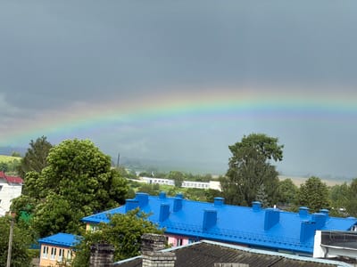 The image captures a post-rain scene with a vibrant rainbow stretching across a cloudy, grey sky. In the foreground, a row of houses with bright blue, tiled roofs is visible, contrasting with the darker tones of the sky. One house features a pink facade with white-trimmed windows. Chimneys made of brick are scattered across the rooftops.

Behind the houses, lush green trees dominate the landscape. The trees are dense and full, indicating a warm season, likely summer, given the foliage. Further in the background, rolling hills are covered in green fields, suggesting a rural or semi-rural setting. Some distant buildings with white walls and windows are faintly visible through a light haze, possibly indicating the edge of a town or a more spread-out settlement.

The overall atmosphere is one of freshness and renewal after a rain shower. The bright colors of the rainbow and the blue roofs stand out against the muted, overcast sky. The scene is peaceful and serene, with no visible people or animals. The light suggests it is daytime, likely late afternoon or early evening, given the intensity of the rainbow and the color of the sky. The location context, Valozhyn, Belarus, places this scene in Eastern Europe. There is no visible text in the image.