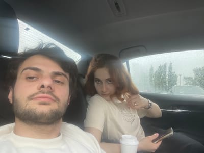 Two people are inside a car. It is raining; raindrops are visible on the car windows. The woman is in a beige shirt and is holding a cup and a phone. The man is in a white sweater. The city and state are Unknown, Georgia.