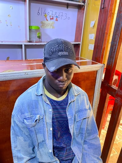 A young person with dark skin is photographed from a slightly low angle, looking directly at the camera. They are wearing a dark baseball cap with a white logo and a light blue denim jacket over a dark blue shirt with a patterned design. The background appears to be indoors, with wooden shelves and a large framed picture or mirror visible behind the person. On a shelf to the left, there is a green-capped plastic container filled with small red beads, and some drawings are taped to the shelf. A handwritten number, "614078730," is also visible on the shelf. The lighting suggests it is daytime. The overall impression is casual and personal. The person's expression is neutral, meeting the viewer's gaze directly. The setting seems to be a shop or a home with display shelves.