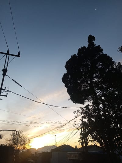 The image depicts an urban or semi-urban street scene at sunrise or sunset in Sa Hti Law (3), Myanmar, characterized by strong backlighting. The sky transitions from a deep blue at the top, featuring a slender crescent moon in the upper right quadrant, to a bright orange and yellow glow at the horizon. Wispy clouds are present in the mid-sky. In the foreground on the left, a utility pole with multiple cross arms and insulators is silhouetted, supporting a complex network of power lines and communication cables that traverse the frame. Some cables appear tangled or show minor debris accumulation. A large, dense tree with irregular foliage dominates the upper right, its form a dark silhouette against the sky. Below the tree and further into the midground, multiple low-rise buildings with varied rooflines and a visible antenna are silhouetted against the bright horizon. Distant mountains form a dark, rugged line along the horizon, obscuring the direct view of the sun. No individuals are visible.