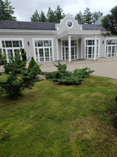 The image depicts a large, modern building with a grey facade and a dark-tiled roof, set against a backdrop of evergreen trees under a cloudy sky. The building features a prominent portico supported by white columns, and large, multi-paned windows. In the foreground, a well-maintained lawn is dotted with several evergreen shrubs. A person wearing a dark hooded jacket and a light-colored skirt is visible standing near the building's entrance, appearing to be looking to the left. The ground in front of the building is paved with light-colored interlocking stones. The scene appears to be located in a semi-rural or suburban area, possibly a public venue or a private residence, within the Старобінскі сельскі Савет region of Belarus, given the context provided.