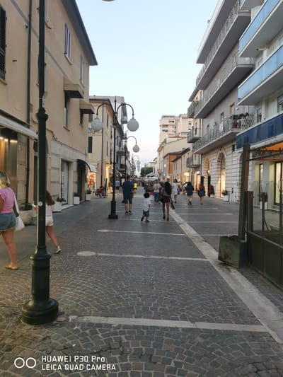 The image depicts a street scene in Follonica, Italy, likely during the evening. A paved pedestrian walkway is lined with buildings on both sides. The buildings are a mix of architectural styles, with some featuring balconies and awnings. Several streetlights with globe-shaped lamps illuminate the area. A number of people are walking along the street, some individually and some in small groups. Some are dressed casually in shorts and t-shirts, suggesting warm weather. The ground is paved with cobblestones. The overall impression is a pleasant, moderately busy street scene.