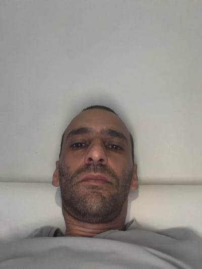 The image is a close-up, low-angle, selfie-style photograph of a man's face. The man has medium-toned skin, short, dark hair, and a beard and stubble. His expression appears neutral. He is wearing a light-colored t-shirt. The background is a plain, light-colored wall or surface, likely indoors. There is no visible indication of the city of Schaerbeek - Schaarbeek, Belgium within the image itself.