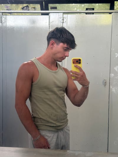 A young man with short, dark hair is taking a mirror selfie in what appears to be a public restroom. He is wearing a light green ribbed tank top and grey sweatpants. He has a silver chain bracelet on his left wrist and is holding a yellow iPhone in his right hand. The restroom has light grey metal stall doors and a light grey tiled floor. There are some stickers on the wall above the stall doors. The overall impression is one of casualness and a focus on physical appearance. The specific location in the city of Δημοτική Ενότητα Προμαχώνος, Greece is not discernible from the image.