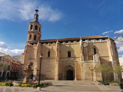 The image depicts the Asunción de Nuestra Señora church in Valdepeñas, Spain. It is a large stone building with a tall bell tower and a tile roof. The church faces the plaza where there is a multi-tiered fountain with yellow flowers. Lampposts are standing along the walls of the building. Some people are visible around the fountain. Another building, possibly an office, is visible on the left with a red exterior. The sky is blue with scattered clouds.