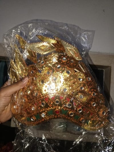 A human hand holds a gold-colored, ornate headdress, which is encased within a transparent plastic bag. The headdress is richly decorated with numerous small, reflective circular sequins or mirrors, alongside larger red and green gem-like embellishments forming a band along its lower edge. Multiple golden, leaf-shaped structures project upwards from the top and sides of the headdress. The hand, visible on the left side of the frame, grips the plastic bag around the base of the headdress. The background consists of a plain, light-colored wall at the upper portion and a dark, indistinct rectangular object, possibly a framed item or furniture, in the lower right background. The specific location in the city of Na Sang, Laos, cannot be determined from the visual information provided.