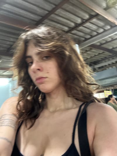 The image presents a close-up, chest-up view of an individual with medium-long wavy brown hair, styled with a middle part, directly facing the camera with a neutral expression. The individual is wearing a black strap top. A tattoo composed of multiple parallel horizontal lines or bands is visible on the left forearm. The background features an overhead structure with a corrugated metal roof supported by exposed beams, suggesting a semi-enclosed outdoor or open-air market-type environment. In the distant right background, a partial view of another person with dark hair and a light-colored top is visible, along with a yellow sign displaying unidentifiable text. The scene is located in the city of Thap Sakae, Thailand.