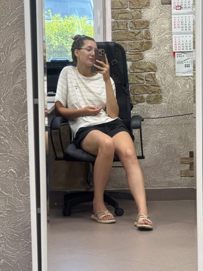 A young woman with her hair in a bun, wearing glasses, a striped t-shirt, and black shorts, sits in a black office chair. She is holding a smartphone in her right hand, taking a selfie in a mirror. Her left hand rests on her lap. She is wearing tan sandals. The background shows a wall with textured wallpaper and a brick pattern, a window with greenery visible outside, and a calendar on the wall. The scene appears to be indoors, possibly in an office or residential space. The floor is tiled. The image is a reflection in a mirror.