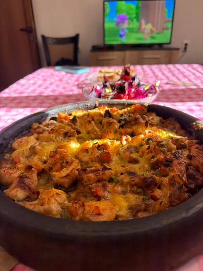 The image displays an indoor scene in Erma Reka, Bulgaria. In the immediate foreground, a dark, shallow earthenware dish (likely a *guvech*) contains a baked meal. The food consists of irregularly cut pieces of chicken, diced carrots, green vegetables, and a layer of melted, golden-brown cheese across the top, showing areas of browning. The dish is situated on a table covered with a pink and white checkered tablecloth.
In the midground, behind the main dish, a clear glass bowl holds an assortment of individually wrapped candies in brightly colored, reflective wrappers, primarily red and purple. Further back on the table, a light blue smartphone is partially visible.
The background reveals a domestic room environment. To the left, a light brown wooden door with a dark handle is closed. A dark wooden chair with a lighter seat back is positioned behind the table. Centered in the background is a flat-screen television resting on a light brown wooden cabinet. The television screen displays a colorful animated scene featuring two cartoon characters, one purple and one orange, in a vibrant green outdoor setting. To the right of the television, a white electrical outlet is visible on the wall. The scene is illuminated by strong, directional light, likely natural sunlight, casting distinct shadows and reflections, particularly on the table and the food. No people are present, and no actions or interactions are depicted.
