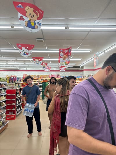 The image depicts the interior of a 7-Eleven convenience store located in Baan Nai Wok, Thailand. The scene is illuminated by overhead fluorescent strip lighting, casting light onto a light-colored tiled floor and white suspended ceiling. Numerous red and white promotional signs featuring cartoon characters such as Hello Kitty and a bear character are suspended from the ceiling.

In the foreground, an individual is partially visible on the right, seen from the chest up, wearing a textured purple t-shirt and a black shoulder strap across his chest. His head is angled downwards.

In the mid-ground, a group of people is visible. To the right, a female with long brown hair, wearing a dark top and a red patterned shawl draped over her shoulders, looks towards the left. She has a ring on her left index finger. Behind her, a male with short dark hair and a beard is partially visible, looking towards the right. To the left, a male with curly dark hair and a beard, wearing a dark blue t-shirt and dark pants, is standing barefoot while holding a six-pack of bottled water. He has white earbuds in his ears and is looking towards the left.

Further in the background, another male individual with dark hair and a beard stands shirtless, wearing dark shorts and carrying a light-colored tote bag over his right shoulder.

The store aisles are stocked with various products. Shelves on the left display beverages, snacks, and multiple jars of Nutella. Deeper into the store, refrigerated sections and other product displays are visible.