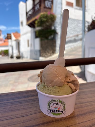 A cup of ice cream with a scoop of pistachio and a scoop of caramel sits on a wooden table in front of a window. The window looks out onto a street in the town of Tejeda, Spain, which is a popular tourist destination on the island of Gran Canaria. There is a white plastic spoon next to the cup. The ice cream is topped with a decorative drizzle of chocolate. The cup has a label that says "Heladeria Nublo Tejeda." The white buildings of the town are visible in the background. The sky is blue and sunny. There is a slight shadow from the window frame on the table.