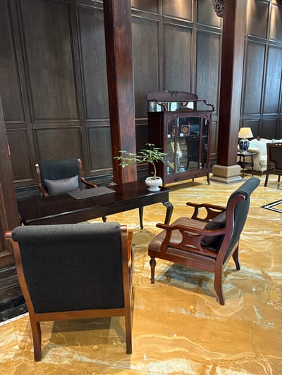 This image captures a static interior scene within a sophisticated lounge or reception area in Tongxiang, China. The environment is defined by dark, vertical wood paneling covering the walls from floor to ceiling, complemented by several large, square-cut dark wooden pillars. The flooring consists of highly polished, light yellowish-brown marble or stone, exhibiting distinctive darker veining and swirling patterns. A small segment of a black and white geometric floor inlay is visible in the far right background.

In the center-left, a dark, polished wooden desk with ornate legs serves as a focal point. On its surface, a small white ceramic pot containing a verdant plant is positioned to the right, and a dark, rectangular mat lies on the left. Three chairs are arranged around this desk: a dark wooden chair with dark gray upholstery and a light gray lumbar cushion is situated behind it; another dark wooden chair with dark gray upholstery occupies the immediate foreground, facing the desk; and a third dark wooden armchair, featuring a brown leather seat, dark gray back cushion, and carved armrests, is positioned to the right of the desk, facing inward.

To the mid-right, a tall, dark wooden display cabinet with glass doors and intricate carvings is visible, containing indistinct items. Further to the right, a cream-colored armchair, a side table, and a table lamp with a white shade and blue patterned ceramic base are partially visible. The scene is devoid of any individuals, actions, or interactions.