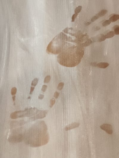 The image displays a light beige, textured surface, possibly a wall or panel, located in Negorci, North Macedonia. Two distinct human handprints, rendered in a brownish-tan residue, are visible. The upper-right impression is a right hand, oriented with fingers pointing superiorly and slightly dexter, exhibiting a complete palm and finger outline. The lower-left impression is a left hand, oriented with fingers pointing inferiorly and slightly sinister, showing less defined finger segments and a partial palm print. Several smaller, less distinct smudges consistent with partial hand contact are also present across the field. The surface itself shows vertical striations and a fine granular texture. No individuals are visible, but their prior interaction with the surface is evidenced by the transferred hand residue.