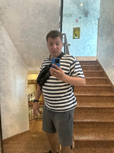 A full-body, medium-shot mirror selfie captures a man standing on a staircase in what appears to be a hotel or apartment building in San Martino Buon Albergo, Italy. The man is wearing a striped t-shirt with navy and white horizontal lines, dark grey shorts, and a black crossbody bag. He is holding a blue smartphone in his right hand, taking the photo. His left arm is resting on his hip. A black watch is visible on his left wrist. The staircase is made of stone or a similar material with a speckled texture in shades of brown and orange. The walls are textured in a pale, mottled greyish-white. In the background, a framed painting with a golden hue hangs on the wall, and a fire safety sign with a red circle and white icon is visible. A metal railing with vertical bars lines the staircase. The lighting suggests an indoor environment, possibly daytime.