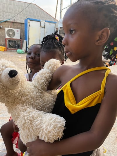 Three young girls are in close proximity, with one holding a large, cream-colored teddy bear. The foremost girl wears a black top with yellow straps and has braided hair adorned with colorful beads. The second girl behind her has braided hair with a single flower accessory. The third girl is partially visible, her expression visible. The background includes a building with an air conditioning unit and a blue and white structure above. The location appears to be Iddah, Nigeria.