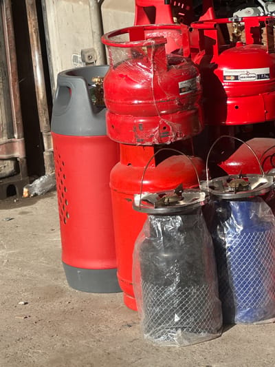This media file displays a collection of red propane gas cylinders, some of which are wrapped in plastic. The cylinders are stacked horizontally, suggesting they are either in storage or awaiting transport. Two cylinders are also visible with blue and black netting over them, respectively, also likely for protection. The scene appears to be outdoors, possibly a marketplace or a storage yard, given the concrete ground and what looks like the edge of a metal structure in the lower left corner, consistent with a bazaar setting in Kara-Suu, Kyrgyzstan. The lighting suggests it is daytime, with shadows cast by the objects. No people are visible in the frame.

One of the red cylinders has a label with visible text. The text includes "ASIA PROPAN" and "7.7kg", indicating the brand and capacity of the cylinder. Another label, partially visible on a different cylinder, contains the text "LPG Cylinder & Pressure Vessels". The overall impression is one of commercial inventory.