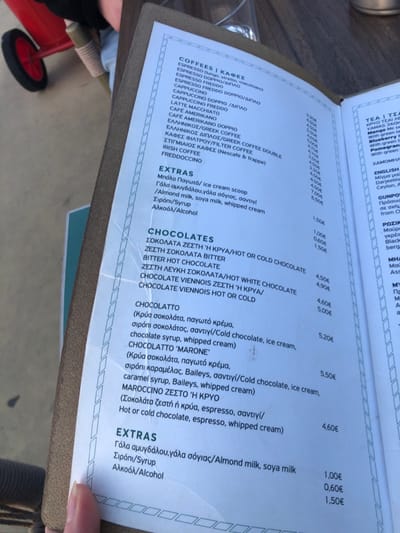 A paper menu on a brown wooden table, listing the prices for coffee and chocolate drinks in a cafe located at 37.929, 23.685 in Palaio Faliro, Greece. The menu is in Greek. The paper is folded in the upper left corner, revealing a red toy wagon partially visible underneath. The camera is pointing directly at the menu.  The table is outdoors. The menu lists the cost of various types of coffee, including espresso, cappuccino, latte, americano, and freddo cappuccino. It also lists the cost of different types of chocolate drinks, including hot chocolate, white chocolate, bitter chocolate, and "Marone." The drinks are available hot or cold. There is also a list of extra ingredients that can be added to the drinks, including milk, cream, ice cream, and whipped cream. The prices range from 1.00€ to 5.50€.