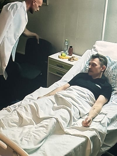 A bald man in a white lab coat is leaning over a man lying in a hospital bed. The man in the bed is wearing a black t-shirt and has an IV drip in his left arm. There is a nightstand next to the bed with a bottle of water and a brown container. The scene appears to be a hospital room.