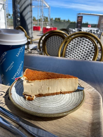 A slice of cheesecake with a crumb topping sits on a decorative plate, with a coffee cup from "Santagloria Coffee & Bakery" to its left, on a wooden table. In the background, outdoor seating with wicker chairs and tables is visible, and beyond that, a stage structure suggests an outdoor event or venue. The lighting indicates it is daytime, and the presence of a clear blue sky points to pleasant weather. The scene is set at an outdoor seating area, likely a cafe or restaurant in Algeciras, Spain, possibly near a public square or event space, given the stage structure.