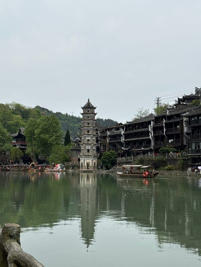 The image shows the reflection of a cityscape in water, likely a river or lake, in Fenghuang, China. The main subjects are the architectural structures, including a distinctive pagoda-like tower and multi-story wooden buildings lining the waterfront. The architecture is traditional Chinese in style with layered roofs and intricate detailing. The environment includes green hills in the background and trees. There is a body of water reflecting the buildings and sky with visible ripples on its surface. The sky is overcast. A boat sits docked at the edge of the building on the waterfront.