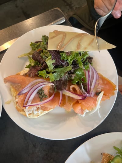 A white plate holds a bagel topped with cream cheese, smoked salmon, capers, and red onion. A mixed greens salad sits beside it. A slice of cheese with a drizzle is pierced by a fork. Another plate with food is partially visible on the upper right. The surface beneath the plate is dark.