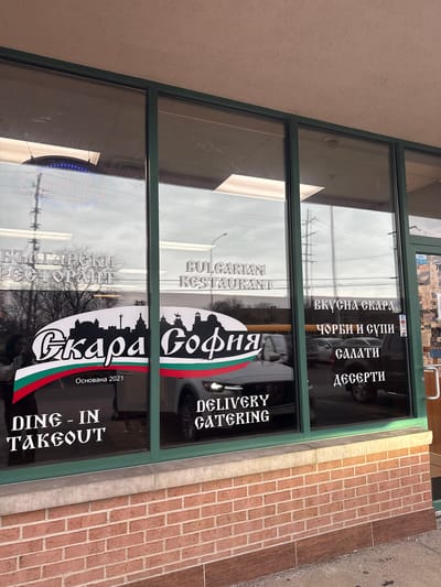 The image shows the storefront of a Bulgarian restaurant in Des Plaines, Illinois, USA. The restaurant's name is "Skara Sofia", indicating that it specializes in grilled dishes ("skara"). The logo incorporates the skyline of Sofia, Bulgaria and the Bulgarian flag. The signage includes the words "Bulgarian Restaurant" in both English and Bulgarian ("БЪЛГАРСКИ РЕСТОРАНТ"). It also specifies "Dine-In/Takeout" and "Delivery/Catering". The menu highlights "delicious grilled dishes" ("ВКУСНА СКАРА"), "soups" ("ЧОРБИ И СУПИ"), "salads" ("САЛАТИ") and "desserts" ("ДЕСЕРТИ").