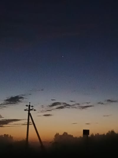 The image depicts a twilight sky over a silhouetted landscape. A tall, V-shaped utility pole with insulators stands prominently on the left side of the frame. To its right, a smaller, dark rectangular object, possibly a signpost, is visible. The horizon is lined with a dense, dark silhouette of trees. The sky transitions from a deep blue at the top to a warm orange and yellow gradient near the horizon, with scattered, dark clouds. A small, faint crescent-shaped celestial body is visible in the upper portion of the sky. The scene appears to be an outdoor, rural or semi-rural environment.