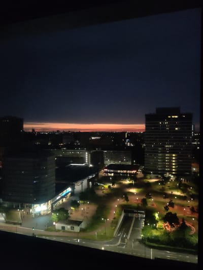 This is a nighttime aerial photograph taken from a high vantage point, likely a building, overlooking a modern urban landscape in Rijswijk, Netherlands. The sky is a deep, dark blue, transitioning to a band of vibrant orange and red along the horizon, indicating the last moments of twilight or an early sunrise.
The foreground shows the edge of a dark, possibly reflective surface, framing the view. In the mid-ground, the scene is dominated by several multi-story buildings. To the right, a tall, rectangular skyscraper stands prominently, its facade illuminated by numerous interior lights visible through the windows, suggesting occupancy. To the left and slightly further back, another large, dark building with a grid-like pattern of windows is visible. In front of these buildings, there are lower structures, some with brightly lit interiors, possibly shops or offices, with illuminated signage.
The ground level reveals a well-lit urban park or plaza area. Numerous streetlights cast a warm glow on pathways, green spaces, and trees. A road with lane markings is visible, and a traffic light shows a green signal. A small, utilitarian building is situated near the road. A car is parked on the road. Further into the park area, there is an entrance to an underground structure, likely a parking garage or tunnel, with visible signage. A lone individual is walking on the pathway near this entrance. The park features various trees and planted areas, some of which are illuminated, creating distinct shadows.
The overall impression is of a well-developed, modern city district, with a mix of residential and commercial structures, and a dedicated green space for public use, all captured during the transition between day and night.
