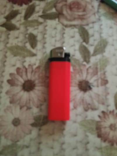 The image shows a bright red disposable lighter with a black cap, placed on a patterned fabric surface. The fabric has a floral design featuring large, muted pink and brown flowers with green leaves. The lighting in the image is diffused, and the focus is on the lighter, although the background pattern is also visible. The setting appears to be indoors, likely on a table or similar surface, given the fabric. There are no people or animals visible in the frame. The image does not provide any information about the city of Zhovti Vody, Ukraine.