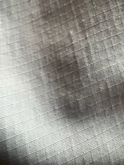 The image displays a close-up, abstract view of a textured fabric. The fabric appears to be a ripstop material, characterized by a grid-like pattern of intersecting threads, creating small squares. The lighting casts shadows and highlights, emphasizing the texture and dimensionality of the weave. The color is predominantly gray, with variations in tone due to the light and shadow. There are no discernible people, objects, or specific environmental features visible that would allow for identification of a location within Mlyniv, Ukraine, or any other specific place. The scene is purely a visual study of textile texture.