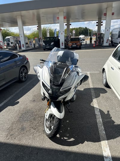 This image captures a sunny day at a gas station in Urvillers, France. The foreground features a white motorcycle parked next to a dark gray car. The motorcycle is positioned slightly left of the frame's center, with its front wheel and fairing prominently displayed. The gray car is to the left of the motorcycle, with its rear visible.
Behind the vehicles, a gas station canopy extends across the width of the image, supported by white pillars. Several gas pumps are visible under the canopy, with numbers indicating different stations. In the background, a black van is parked at one of the pumps, and an orange car is situated further down. A white commercial van is parked to the right, and a person is seen walking near the orange car. The sky is clear with scattered clouds, and trees line the distant horizon. The ground is paved asphalt, marked with white lines indicating parking spaces.