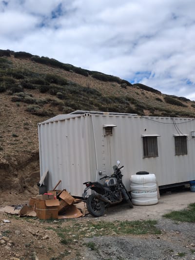 The image depicts an outdoor scene featuring a white shipping container modified into a structure, likely a temporary building or shelter. The container is situated on a dirt and gravel ground against a backdrop of a barren, rocky hillside covered with sparse vegetation. In front of the container, there is a pile of discarded cardboard boxes, some open and others stacked. To the right of the boxes, a grey motorcycle with a license plate is parked. Next to the motorcycle, a large white plastic water tank is positioned. The sky above is partly cloudy, with patches of blue visible between white clouds. The overall impression is one of a functional, utilitarian setting in a remote or undeveloped area. There are no people visible in the image. The location is identified as Dhar Chharp Samba, India, suggesting a rural or mountainous environment within that region.