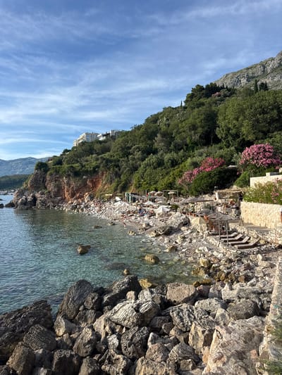This image depicts a picturesque coastal scene in Rijeka Reževići, Montenegro. The foreground is dominated by large, weathered rocks of varying sizes and textures, bordering a crystal-clear, turquoise sea. The water is calm, with small waves gently lapping at the shore.

The middle ground features a rocky beach dotted with numerous sun loungers and straw umbrellas, indicating a recreational area. Several people are visible, relaxing on the loungers or walking along the beach. Behind the beach, a stone wall and terraces rise up the hillside, adorned with lush green vegetation and vibrant pink oleander flowers. Further up the incline, dense green trees cover the slope, leading to the base of a rugged, grey mountain.

In the upper portion of the image, a modern, multi-story white building with large windows is perched on the hillside amidst the greenery. The sky above is a clear blue with wispy white clouds, suggesting fair weather. The overall impression is one of a serene and beautiful seaside resort.