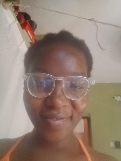 A woman with dark skin and short black hair is seen from a low angle, wearing clear-framed glasses. She is smiling slightly. An orange tank top is visible. The background includes a white wall with some items hanging, possibly toys and a wire. A green wall with a doorway is also visible in the background, creating an indoor setting. The glasses have noticeable glare on the lenses.