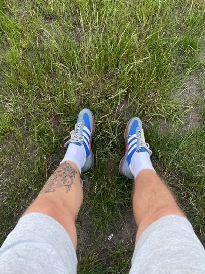 A person's legs are shown from a top-down perspective, standing on a patch of grass in what appears to be a rural or park area. The individual is wearing light grey athletic shorts, white socks, and blue and grey Adidas sneakers with white stripes. A tattoo is visible on the left calf, depicting intricate designs, including what looks like a face or figure. The background consists of green grass and dark soil, suggesting an outdoor setting. The specific location within Азершчынскі сельскі Савет, Belarus, is not determinable from the visual information alone.