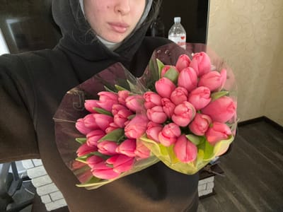 The image displays a female individual holding two large bouquets of vibrant pink tulips in an indoor setting located in Slavyansk-na-Kubani, Russia. The individual is partially visible, cropped from the nose downwards, wearing a black hooded sweatshirt with the hood up, over what appears to be a white turtleneck or high-collared shirt. Both bouquets are wrapped in clear plastic cellophane, with the tulip buds predominantly closed. The tulips exhibit a uniform pink hue. In the background, a white vertical structure and a decorative white brick-patterned element are visible to the left. To the upper right, a clear plastic water bottle with a white cap, featuring a partially visible Cyrillic label, rests on a surface against a light-colored patterned wallpaper. The floor is dark and reflective.
