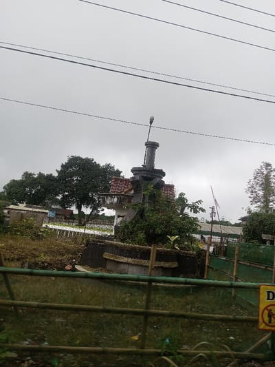 The image depicts a static outdoor scene in Eheliyagoda, Sri Lanka, under a uniformly overcast sky. Central to the composition is a weathered, multi-component concrete monument or structure, approximately two meters in height, constructed with stacked geometric shapes. It features a broad, dark-colored base with a rounded edge, supporting a narrower, tiered structure with a reddish-brown tiled roof section on its left side. A prominent gray cylindrical column rises from the top, surmounted by a finial-like element that appears to be a street light fixture, angled slightly upwards to the left. Dense green foliage partially obscures the base of the monument.
The foreground consists of unkempt grassy terrain with scattered refuse. A horizontal light-colored wooden or bamboo fence delineates the immediate foreground. On the far right, a yellow warning sign with a red circular 'no entry' symbol featuring a human figure is partially visible, attached to this fence. Beyond the monument, in the middle ground, are various utilitarian structures, including what appears to be a pond or waterway with white parallel structures possibly related to aquaculture, and a dark green, mature tree dominating the left background. Other distant buildings with red-tiled roofs are faintly visible. The upper portion of the frame is crossed by several taut, dark electrical or utility wires running horizontally. No human figures or active interactions are visible.