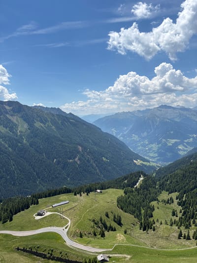 The image captures a high-altitude landscape in St. Leonhard in Passeier - San Leonardo in Passiria, Italy, under a bright blue sky with scattered white cumulus clouds. The foreground features a winding asphalt road cutting through lush green rolling hills dotted with scattered evergreen trees. A small building with a dark roof sits beside the road, and further up the hill, a larger, long, barn-like structure is visible. In the mid-ground, the hills give way to densely forested mountains, their slopes covered in dark green coniferous trees. The background reveals a majestic panorama of rugged mountain ranges receding into the distance, with a deep valley cutting through them, revealing hints of human settlements with lighter green patches. The overall scene evokes a sense of vastness and natural beauty.