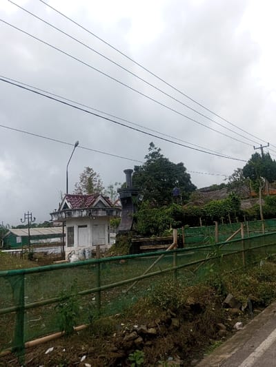 An outdoor scene in Sarwahi Mal., India, depicts a semi-rural environment under an overcast, grey sky. Multiple black utility lines traverse the upper portion of the frame. In the immediate foreground, a dirt and rock embankment with sparse weeds transitions into a narrow, dark grey paved surface, likely a road or path, along the lower right edge.
A green mesh fence, supported by bamboo poles, stretches horizontally across the mid-ground, separating the foreground embankment from a grassy area. Beyond this fence, to the left of center, stands a small, white, single-story structure with a red-tiled hip roof featuring decorative gables. Its front presents a closed white wooden door and a window. Several white sacks are placed at its base. A black streetlamp post with a single luminaire extends upward to the left of this building.
To the right of the white building, a dark, vertical monument or pillar structure, possibly stone or concrete, is partially visible, obscured by dense green foliage. Further into the background, a large area of lush green trees and shrubs dominates, within which a blue water tank on a metal stand is discernible. Parts of residential structures with tiled roofs are faintly visible amidst the dense vegetation on the right. In the far left background, a green and white building, potentially a greenhouse or agricultural structure, is visible, along with another decorative metal utility pole. No individuals or animals are present.